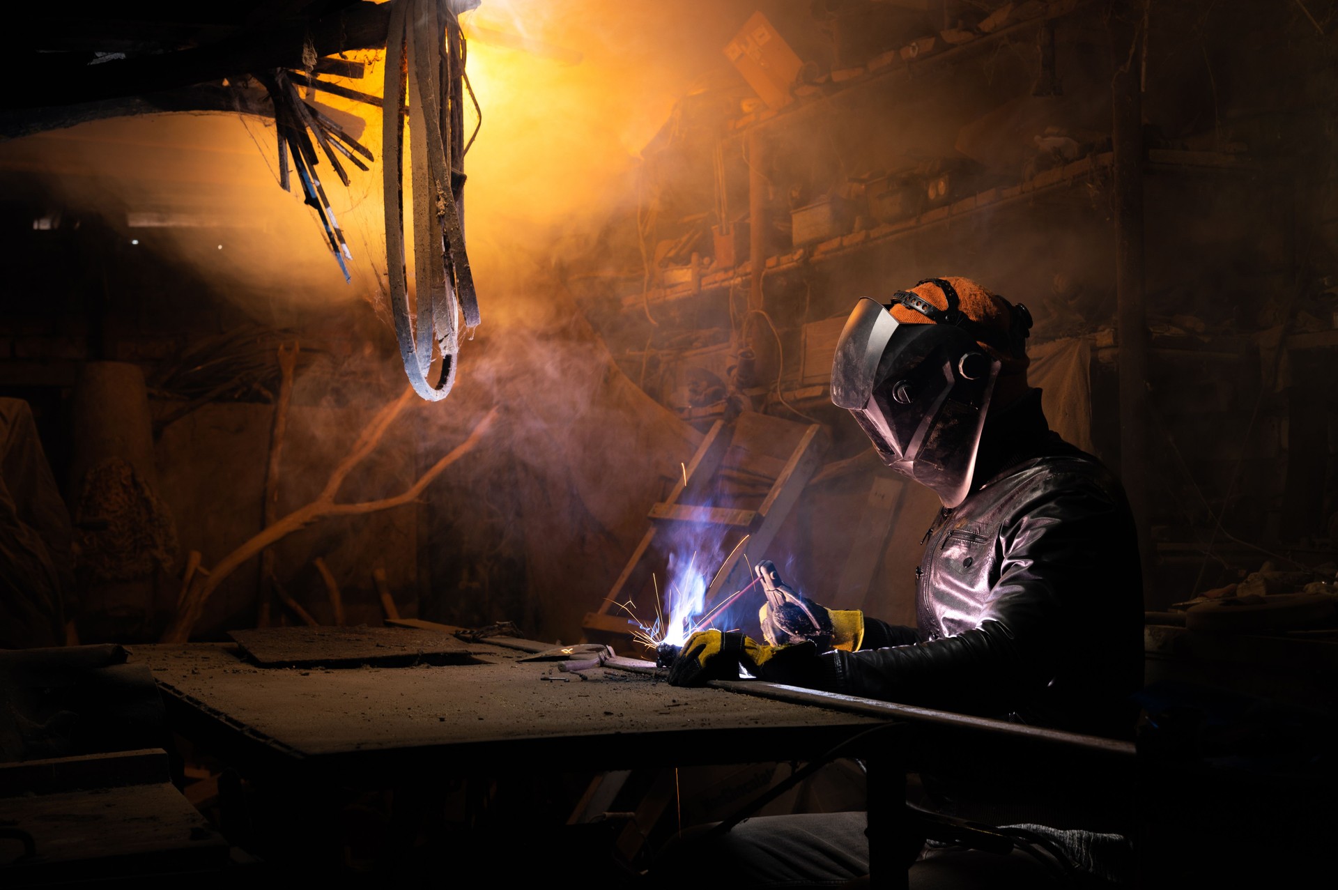 A welder in a protective mask for metal welding and protective gloves in a leather jacket performs welding work in a home garage. Sparks, hot flame from welding.